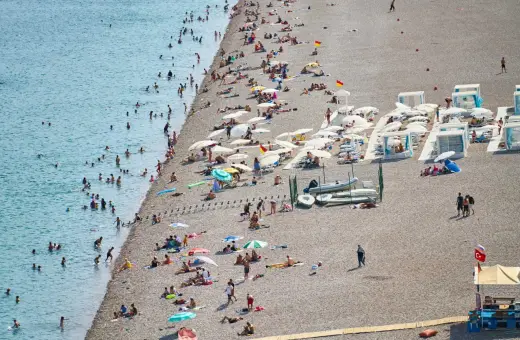 Tłum ludzi odpoczywa na kamienistej plaży w Finike. Niektórzy kąpią się w morzu, inni opalają się pod parasolami.