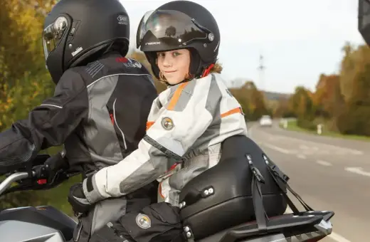 Uśmiechnięte dziecko na motocyklu, siedzące za kierowcą, w kombinezonie i kasku, gotowe na przygodę.