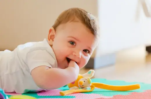 Uśmiechnięte 11 miesięczne dziecko leży na macie edukacyjnej, gryząc paluszek. Obok pluszowa zabawka.