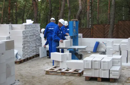 Budowa trwa! Pracownicy w niebieskich kombinezonach i białych kaskach układają bloczki ytong. Na paletach widać, że ytong 12 to spora ilość materiału budowlanego.