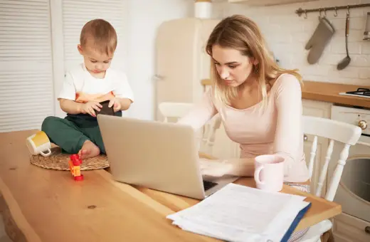 Młoda mama pracuje na laptopie, podczas gdy jej małe dziecko bawi się obok. Czy na urlopie macierzyńskim można pracować? Tak, często jest to możliwe.
