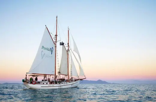 Żaglowiec "Providence" płynie po spokojnych wodach, zbliżając się do Whitehaven Beach. Zachód słońca maluje niebo pastelowymi barwami.