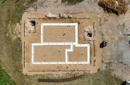 Widok z lotu ptaka na plac budowy. Widać fundamenty domu, piasek, drewno i wykopy pod studnie fundamentowe.
