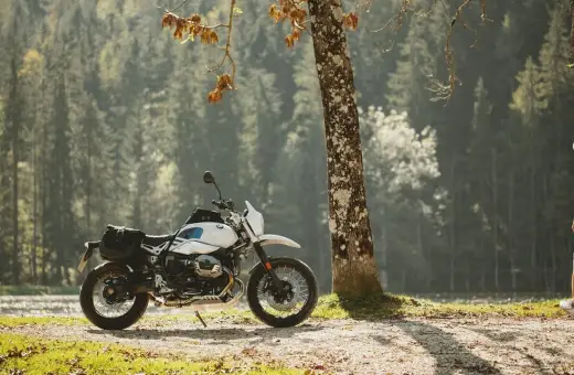 Biały motocykl z sakwami, zaparkowany przy drzewie w lesie. Idealny do testów w rankingu opon motocyklowych.