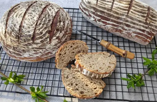 Dwa bochenki chleba żytniego na zakwasie, jeden okrągły, drugi podłużny, z kilkoma kromkami na ruszcie.