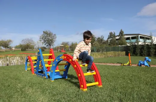 Kolorowy plac zabaw w ogrodzie to świetne atrakcje na urodziny dziecka. Chłopiec wspina się po zjeżdżalni, a obok stoi jeździk i konik na biegunach.