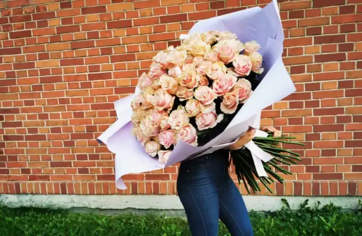 Ogromny bukiet 108 róż w odcieniach różu i brzoskwini, owinięty w fioletowy papier, trzymany przez osobę w dżinsach na tle ceglanej ściany.