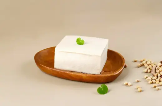 Kostka białego tofu na drewnianej miseczce, ozdobiona listkiem. Obok garść ziaren soi. To właśnie co to tofu – produkt z soi.
