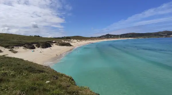 Vista panorámica de la playa de Santa Comba, con arena dorada, aguas turquesas y colinas verdes. Algunas personas disfrutan del sol y el mar.