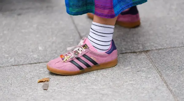 Różowe buty sportowe Adidas Gazelle z czarnymi paskami i charmsami, białe skarpetki w paski. W tle widać drugie, podobne buty w innym kolorze.