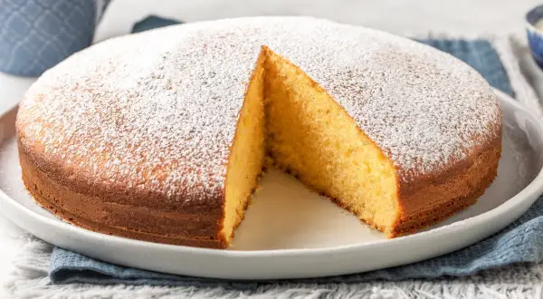 Torta yogurt greco soffice e dorata, cosparsa di zucchero a velo. Una fetta è stata tolta, rivelando l'interno profumato.