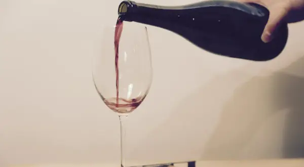 Ein Weinglas wird mit Rotwein gefüllt. Eine Hand hält die dunkle Flasche, während der Wein in einem dünnen Strahl ins Glas fließt.