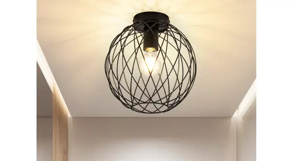 Nowoczesna lampa sufitowa z czarną, drucianą kulą i widoczną żarówką, tworząca ciekawe cienie na suficie.