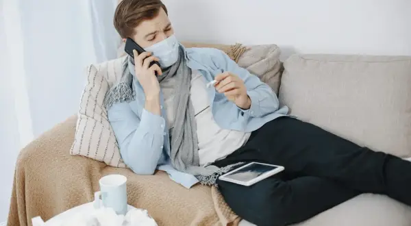 Chory mężczyzna w masce i szaliku leży na kanapie, rozmawiając przez telefon i trzymając termometr.
