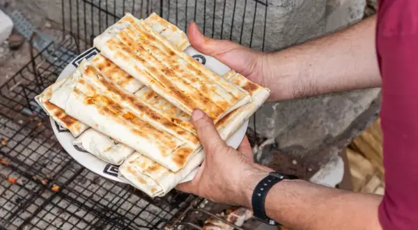 Talerz z grillowanymi wrapami kebab, trzymany przez ręce na tle rusztu i kamieni.