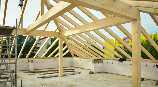 Struttura in legno per la realizzazione di tetti, con travi a vista e muri in blocchi di cemento. In sottofondo, attrezzature da cantiere.