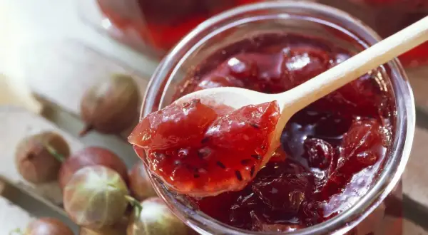 Ein Holzlöffel mit leckerer Stachelbeermarmelade ragt aus einem Glas. Frische Stachelbeeren liegen daneben.