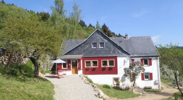 Maison de campagne avec terrasse et parasol, idéale pour un camping à la ferme dans les Vosges. Le soleil brille sur ce paysage verdoyant.