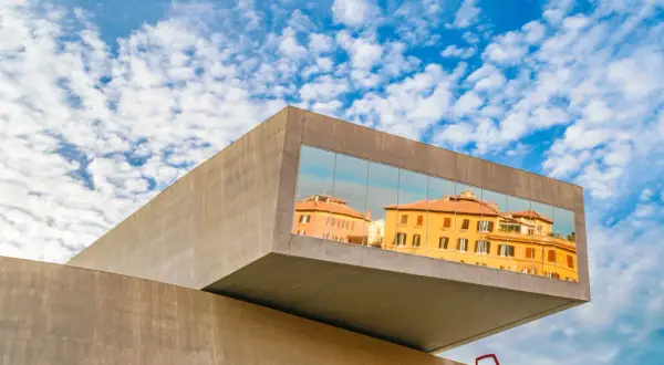 Il MAXXI - Museo Nazionale delle Arti del XXI Secolo riflette edifici storici romani sotto un cielo azzurro e nuvoloso.