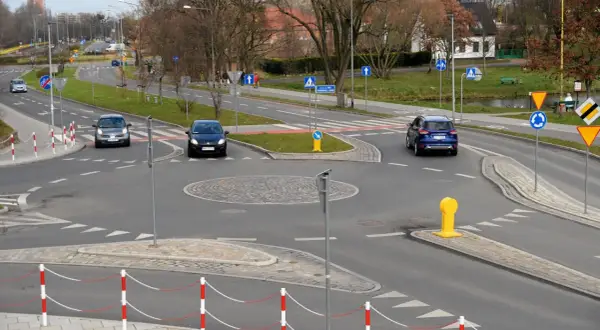 Samochody zbliżają się do ronda. Widoczny jest kierunkowskaz na rondzie, a także znaki drogowe i bariery.