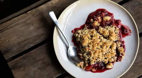 Ein Teller mit warmem Pflaumen Crumble, dazu ein Löffel auf rustikalen Holzdielen.