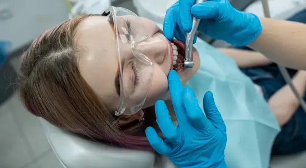 Pacjentka w okularach ochronnych przechodzi leczenie kanałowe. Dentysta w niebieskich rękawiczkach używa wiertła, by ulżyć w bólu zęba.