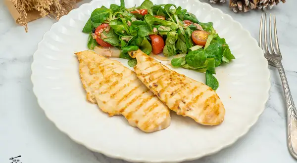 Dos pechugas de pollo a la plancha con marcas de parrilla, servidas con una ensalada fresca de hojas verdes y tomates.