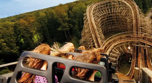 Fahrt mit einer Holzachterbahn, Menschen mit wehenden Haaren, im Hintergrund Wald und Himmel.