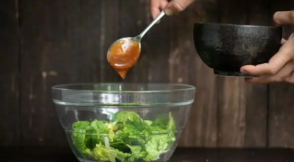 Ein Löffel mit braunem Salatdressing tropft in eine Schüssel mit knackigem Salat.