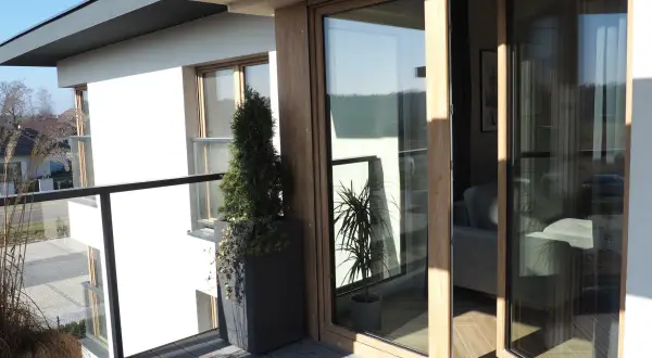 Nowoczesne okna dzielone z drewnianymi ramami otwierają widok na balkon z roślinnością i wnętrze domu.