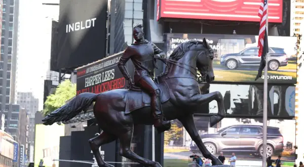 A bronze statue of a person on horseback stands in Times Square, with billboards for Inglot and Harper Lee's "To Kill a Mockingbird" in the background.