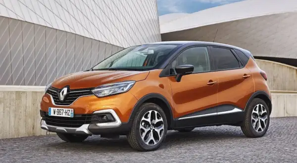 Pomarańczowy Renault Captur 2016 z czarnym dachem zaparkowany na brukowanej nawierzchni przed nowoczesnym budynkiem.