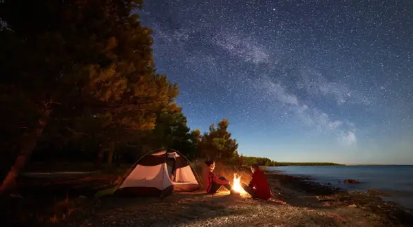 Nocne biwakowanie pod rozgwieżdżonym niebem. Dwoje ludzi siedzi przy ognisku obok namiotu z materiału namiotowego, na kamienistej plaży.