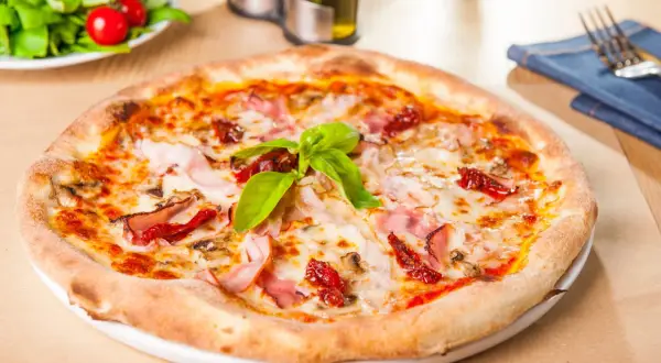 Pizza garnie de jambon, champignons et tomates séchées, sur une pâte à pizza moelleuse. Une feuille de basilic frais couronne le tout.