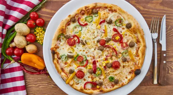 Apetyczna pizza z warzywami i mięsem na talerzu, obok sztućce i składniki.