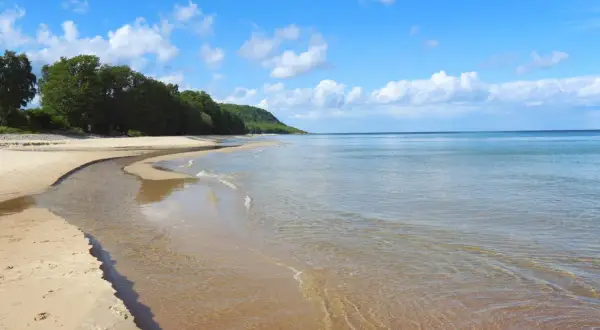 En stilla stenshuvud strand med klart vatten som möter en sandig kustlinje. Grönska och kullar syns i bakgrunden under en blå himmel med vita moln.