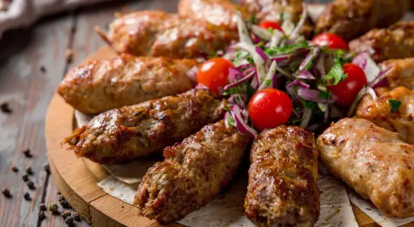 Kebab ormiański na drewnianej desce, podany z sałatką z pomidorków i cebuli.