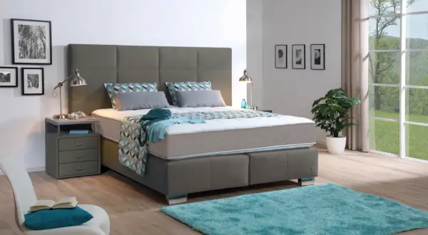 Ein modernes Schlafzimmer mit einem grauen Boxspringbett, das mit einer geometrisch gemusterten Decke und Kissen bezogen ist. Ein türkiser Teppich liegt davor.