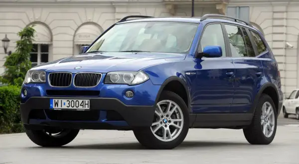 Niebieskie BMW X3 2005 z polskimi tablicami rejestracyjnymi WI 3004H zaparkowane przed budynkiem.