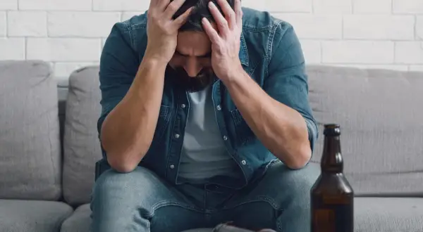 Man met handen in het haar, zittend op een bank met een fles bier. Dit illustreert de definitie van verslaving: een dwangmatige behoefte.