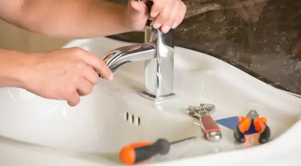 Ręce majsterkowicza naprawiają cieknący kran dwuuchwytowy. Obok leżą klucz, śrubokręt i kombinerki.