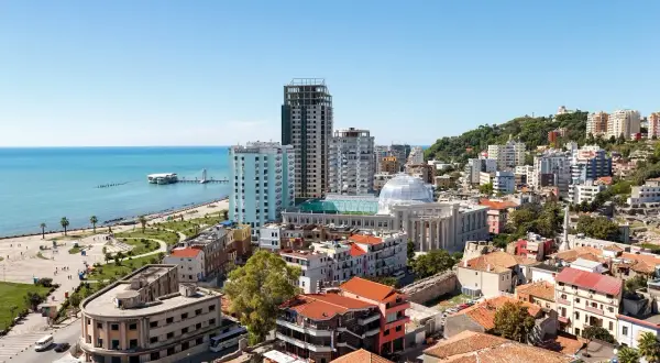 Widok na miasto Durres z plażą w tle. Widać morze, promenadę, budynki mieszkalne i hotelowe oraz wzgórze porośnięte zielenią.