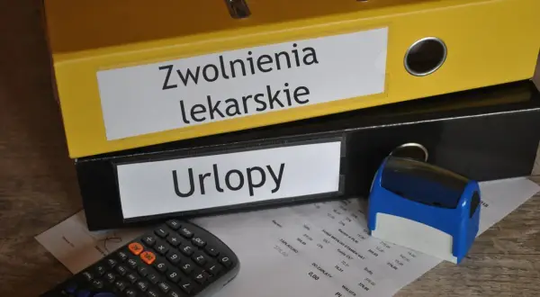 Zółty segregator "Zwolnienia lekarskie" i czarny "Urlopy" obok kalkulatora i pieczątki. Dokumenty na biurku sugerują, że kod a na zwolnieniu jest w trakcie rozliczeń.