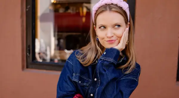 Uśmiechnięta, clean girl w dżinsowej kurtce i różowej opasce na głowie, opiera policzek na dłoni.