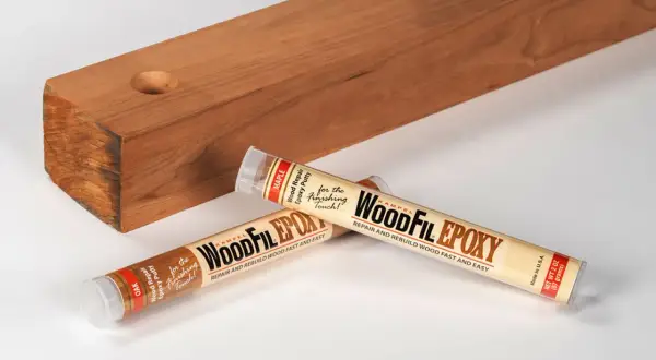 Two tubes of WoodFil Epoxy, a wood repair putty, lie next to a wooden plank with a hole, demonstrating how to use wood filler for quick repairs.