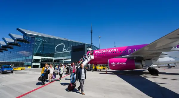 Pasażerowie wsiadają do różowego samolotu Wizz Air na płycie lotniska. Nowoczesny terminal lotniczy w tle.
