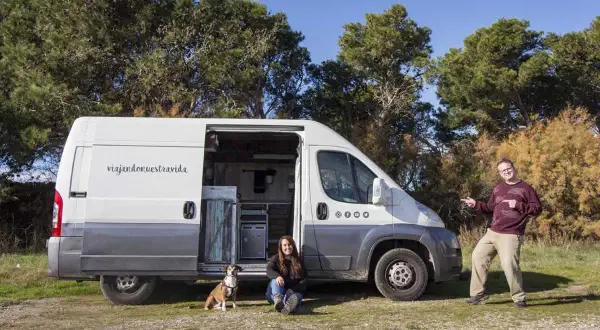 Pareja y perro posan junto a su furgón vivienda, listos para viajar.