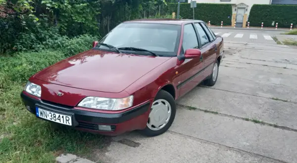 Czerwony Daewoo Espero z tablicą rejestracyjną WN-3841R zaparkowany przy zarośniętym poboczu i betonowej drodze.