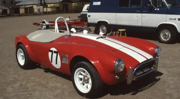 Röd sportbil, en kungens AC Cobra med nummer 77, redo för race. Bakom skymtar en blåvit skåpbil.