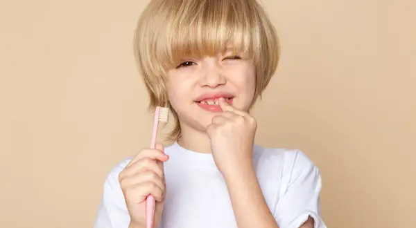 Chłopiec z blond włosami i różową szczoteczką w dłoni pokazuje palcem bolące dziąsło, być może objaw wirusowego zapalenia dziąseł u dziecka.